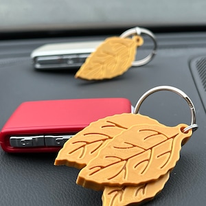 May include: Two gold-colored leaf-shaped keychains with silver key rings. The keychains are on a black surface.