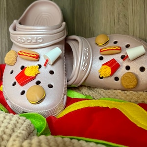 May include: A pair of pink Crocs decorated with various food charms, including a hot dog, french fries, a hamburger, and a milkshake. The charms are attached to the shoes with holes.