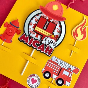 May include: Firefighter themed cake topper set with a red fire truck, fire hydrant, a Dalmatian puppy, a flame, and a red circle with the number 4 and the name Micah.