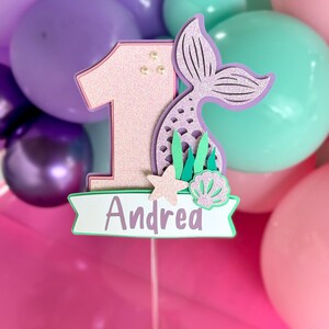 May include: A glittery pink number one cake topper featuring a purple mermaid tail, a small starfish, and a seashell. A white banner at the bottom displays the name Andrea in purple text. The topper is mounted on a thin stick for placement.