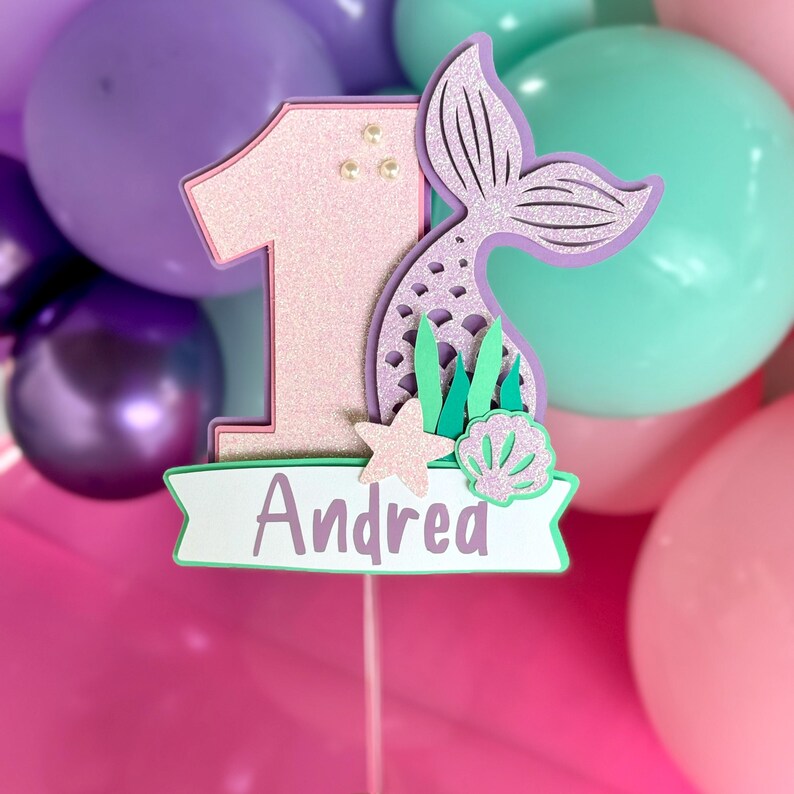 May include: A glittery pink number one cake topper featuring a purple mermaid tail, a small starfish, and a seashell. A white banner at the bottom displays the name Andrea in purple text. The topper is mounted on a thin stick for placement.