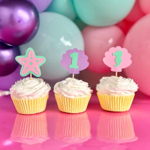 May include: Three cupcakes in yellow paper liners topped with white frosting and decorative cardstock toppers. The toppers feature a pink starfish, a purple shell with the number one, and a pink shell with a green seahorse silhouette.