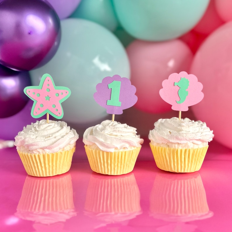 May include: Three cupcakes in yellow paper liners topped with white frosting and decorative cardstock toppers. The toppers feature a pink starfish, a purple shell with the number one, and a pink shell with a green seahorse silhouette.