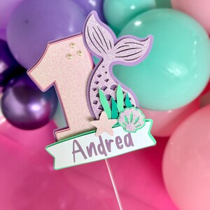 May include: A cake topper featuring a glittery light pink number one, a lavender mermaid tail, a small beige starfish, and a light green seashell. A white banner at the bottom displays the name Andrea in purple text, all mounted on a thin stick.
