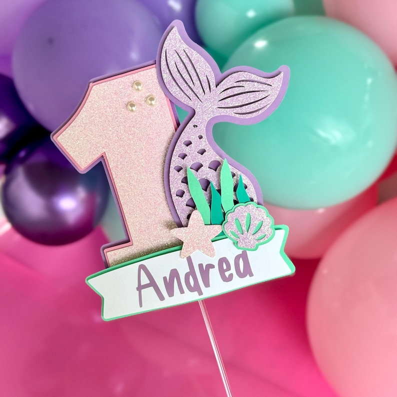 May include: A cake topper featuring a glittery light pink number one, a lavender mermaid tail, a small beige starfish, and a light green seashell. A white banner at the bottom displays the name Andrea in purple text, all mounted on a thin stick.