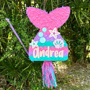 May include: A pink, purple, and green mermaid tail pinata with the name "Andrea" written on it. The pinata has a pink tail fin, purple and green scales, and a blue and white seashell design. It has a pink and purple tassel.