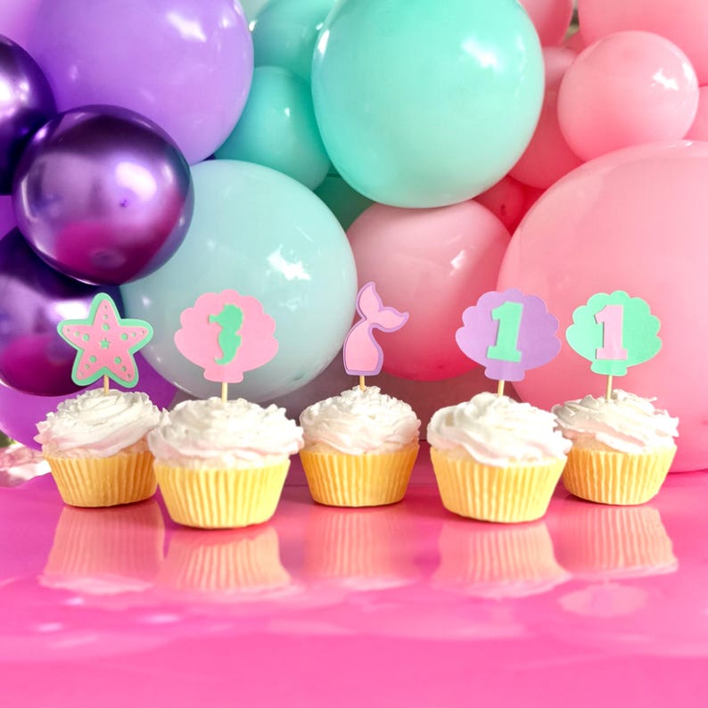 May include: Five cupcakes with white frosting topped with paper decorations including a pink starfish, a green seahorse in a shell, a pink mermaid tail, and two shells featuring the number one, set against a background of purple, teal, and pink balloons.