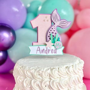 May include: A glittery pink number one cake topper featuring a purple mermaid tail, a small starfish, and a seashell. A white banner at the base displays the name Andrea in purple text. The topper is positioned on a white frosted cake.