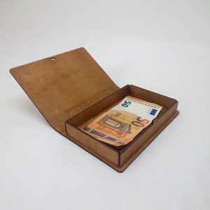 Puede incluir: Una caja de madera marrón con forma de libro con un billete de 50 euros en su interior. La caja está abierta y el billete es visible.