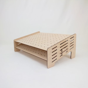 Puede incluir: Una estantería de madera de dos niveles con una superficie perforada y un panel lateral con un diseño recortado. La estantería está hecha de madera clara y tiene un acabado natural.