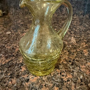 May include: A yellow-green glass pitcher featuring a distinct crackle texture throughout its surface. The vessel has a wide, rounded base that tapers into a narrow neck, a small pouring spout, and a curved handle attached to the side.