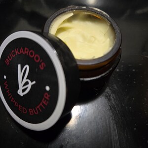 May include: A small amber glass jar filled with pale yellow whipped butter. The black lid is resting beside the jar, featuring a white circular border, the text "BUCKAROO'S WHIPPED BUTTER" in red, and a large white letter "B" in the center.