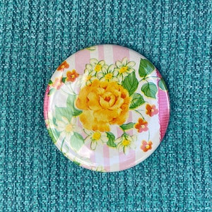 May include: Round floral button with a yellow rose and other flowers on a pink and white striped background. The button has a glossy finish and is surrounded by a pink border. The button is set against a teal textured surface.