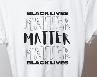 May include: White t-shirt with the text "BLACK LIVES MATTER MATTER MATTER BLACK LIVES" in black, handwritten-style font. The shirt is hanging on a wooden hanger.