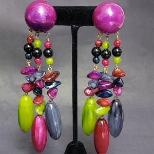 May include: A pair of long, dangling earrings with a large, round, purple top. The earrings feature a variety of colorful beads, including green, red, pink, blue, and black.