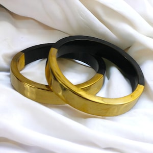 May include: Two circular bangle bracelets featuring a two-tone design. Each bangle is constructed with a smooth, matte black interior and a polished, reflective gold-tone exterior. The bracelets have a wide, flat profile and a solid, rigid structure.