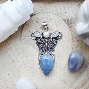 Peut inclure: Un pendentif en argent arborant un motif de papillon aux ailes en filigrane complexe, surmonté de trois petites pierres blanches rondes et ovales, et terminé par une grande pierre bleue lisse en forme de goutte, suspendu à une bélière en argent.