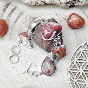 Può includere: Un bracciale in argento con pietre di diaspro rosso e marrone di varie forme. Il bracciale è composto da più segmenti collegati, ognuno con una pietra di forma diversa. Le pietre sono incastonate in castoni d'argento.