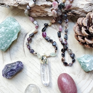 Peut inclure: Un collier de pierres précieuses avec un pendentif en quartz clair et diverses perles colorées. Le collier est présenté avec des pierres précieuses brutes et une pomme de pin sur une surface en bois. Les perles sont dans des tons de rose, de violet et de noir.