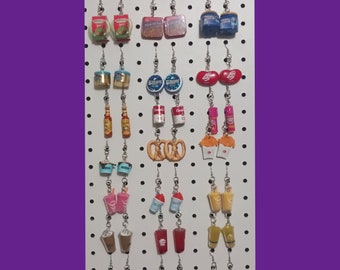 May include: A white pegboard with 20 pairs of dangle earrings featuring various food and drink themed charms. The charms include:  Wheaties cereal boxes,  a can of Spam,  a can of  'Dinty Moore' soup,  a can of  'Campbells' soup,  a bag of  'Jelly Belly' jelly beans,  a bottle of hot sauce,  a pretzel,  a cup of ice cream,  a cup of  'Slurpee',  a cup of  'Jamba Juice',  a cup of coffee,  a cup of  'Red Bull',  a bottle of water,  and a cup of boba tea.