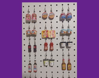 May include: A white pegboard with various food-themed earrings. The earrings are made of miniature food items, such as cereal boxes, cans of soup, and bottles of hot sauce. The earrings are arranged in rows on the pegboard.