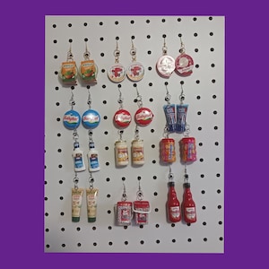 May include: A white pegboard with various food-themed earrings. The earrings are made of polymer clay and feature popular food brands like Babybel, Wheaties, and Red Hot.