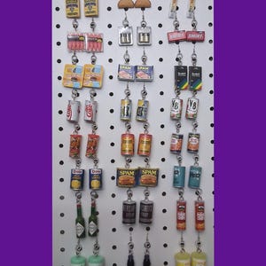 May include: A collection of 20 pairs of earrings featuring miniature food and beverage items. The earrings are displayed on a white pegboard with silver hooks. Some of the items include: Spam, Coke, Dole pineapple, Hershey's syrup, Wet Ones, and Planters peanuts.