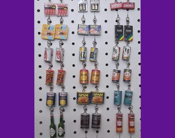 May include: A collection of 20 pairs of earrings featuring miniature food and beverage items. The earrings are displayed on a white pegboard with silver hooks. Some of the items include: Spam, Coke, Dole pineapple, Hershey's syrup, Wet Ones, and Planters peanuts.