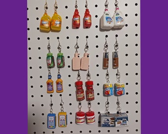 Peut inclure: Boucles d'oreilles miniatures représentant des aliments et des boissons, notamment des bouteilles de jus, des cartons de lait et des pots d'épices, suspendues à un panneau perforé blanc. Les boucles d'oreilles sont fixées à des crochets argentés.