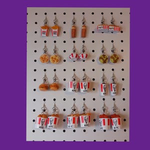 May include: A collection of novelty earrings featuring miniature food items. The earrings include fries, chicken, burgers, and KFC buckets. The earrings are attached to silver-colored hooks and displayed on a white pegboard with black dots.