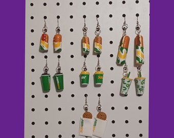 May include: A white pegboard with a variety of Subway and Jimmy John's themed earrings. The earrings are made of polymer clay and feature different food items, including sandwiches, drinks, and cookies.
