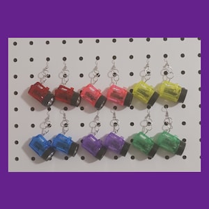May include: A set of 10 colorful, miniature flashlight earrings. The earrings are made of clear plastic and have a black metal hook. The flashlights are in various colors, including red, pink, yellow, blue, green, and purple.