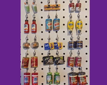 May include: A collection of novelty earrings featuring miniature food and beverage items. The earrings include tiny versions of condiments, snacks, and canned goods, such as ketchup, peanut butter, and soup cans. The earrings are displayed on a white pegboard.