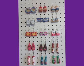 May include: A pegboard with various food-themed earrings. The earrings are made of polymer clay and feature different food items, such as cans of soup, bottles of ketchup, and boxes of cereal.