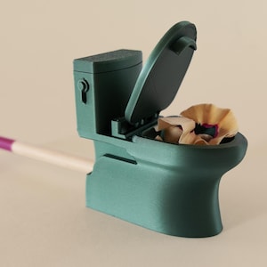 Puede incluir: Un sacapuntas de color verde azulado con forma de inodoro, con la tapa abierta y virutas de lápiz en su interior. Un lápiz con una goma de borrar morada está insertado. El fondo es de color beige liso.