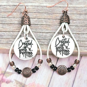 May include: A pair of dangle earrings featuring white leather loops, copper-toned hooks, and textured metal caps. Each earring holds a round silver-toned charm stamped with the black text Created with a purpose, accented by black and copper beads.