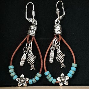 May include: A pair of dangle earrings featuring a brown leather hoop, turquoise-colored beads, and silver-toned metal charms. Each earring includes a small fish charm, a loaf-shaped charm, and a five-petaled flower charm at the base.