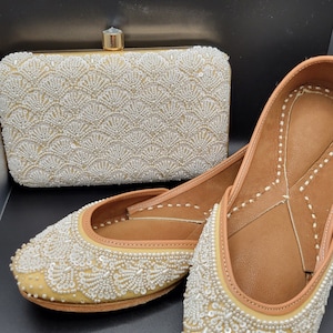 May include: A pair of beige flat shoes with white pearl embellishments and a matching clutch bag. The shoes and bag are decorated with a pattern of white pearls.