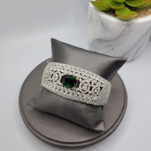 May include: A silver-toned bracelet with a rectangular emerald-green center stone and numerous clear, sparkling accent stones. The bracelet is displayed on a gray satin pillow and a gray circular base, with a succulent plant in the background.
