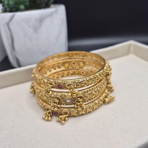 May include: A set of three gold-toned bangles featuring intricate embossed floral patterns, small dangling charms, and embedded circular red and clear stones. The metal surface has a matte finish with detailed engravings throughout the bands.