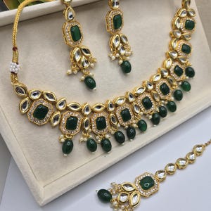 Peut inclure: Un ensemble de bijoux doré comprenant un collier, des boucles d'oreilles et un bijou de tête. L'ensemble comprend des pierres carrées vert émeraude, de petites perles blanches et des cristaux transparents. Le collier et les boucles d'oreilles ont un motif floral.