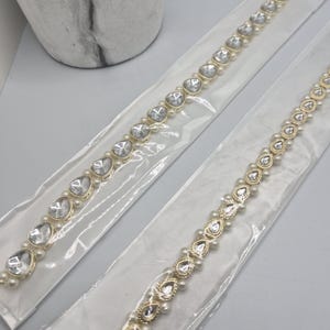 May include: Two gold-toned, delicate headbands with clear, heart-shaped stones and small pearls. The headbands are in clear plastic packaging, with a marble-patterned vase in the background. The jewelry is likely for a special occasion.
