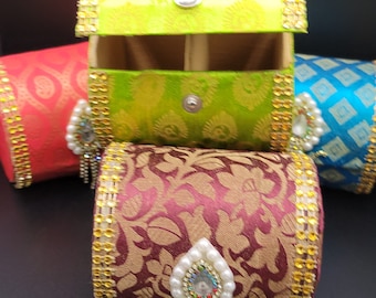 May include: A selection of decorative, cylindrical boxes in shades of red, blue, green, and maroon. Each box is embellished with gold trim and a decorative jewel. The boxes feature a floral pattern.