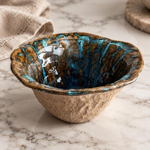 May include: A small ceramic bowl with a rough, textured beige exterior and a glossy, vibrant interior featuring a blend of deep brown and bright turquoise blue glaze. The rim is uneven and organic in shape, showcasing a hand-crafted appearance.