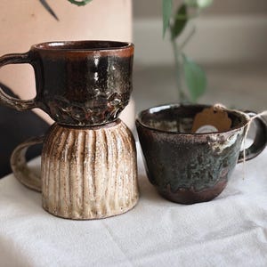 May include: Three ceramic mugs with a dark, glossy glaze and textured surfaces. One mug features vertical ribbing on the lower half, while the others have a smooth, mottled finish. Each mug has a sturdy handle and a rustic, handcrafted appearance.