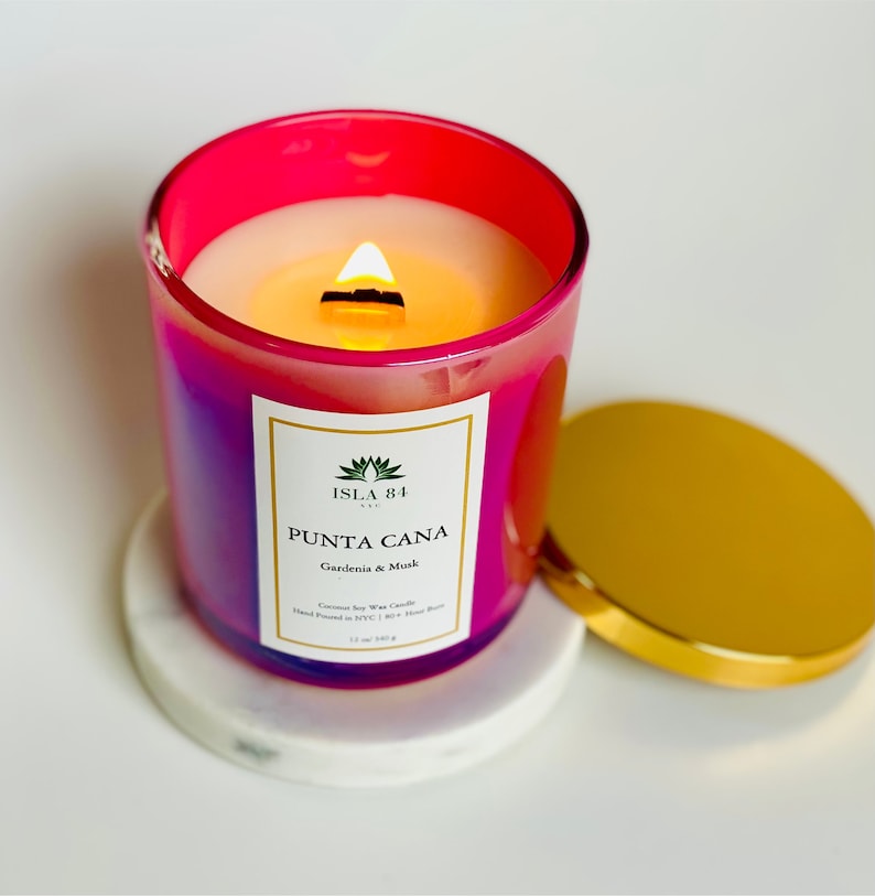 K&ouml;nnte beinhalten: Eine brennende, leuchtend rosa und lila Glas-Kerze mit goldenem Deckel, die auf einem wei&szlig;en Marmor-Untersetzer steht. Die Kerze hat ein Etikett mit der Aufschrift "Punta Cana" und "Gardenia & Musk". Die Kerze ist 340 g.