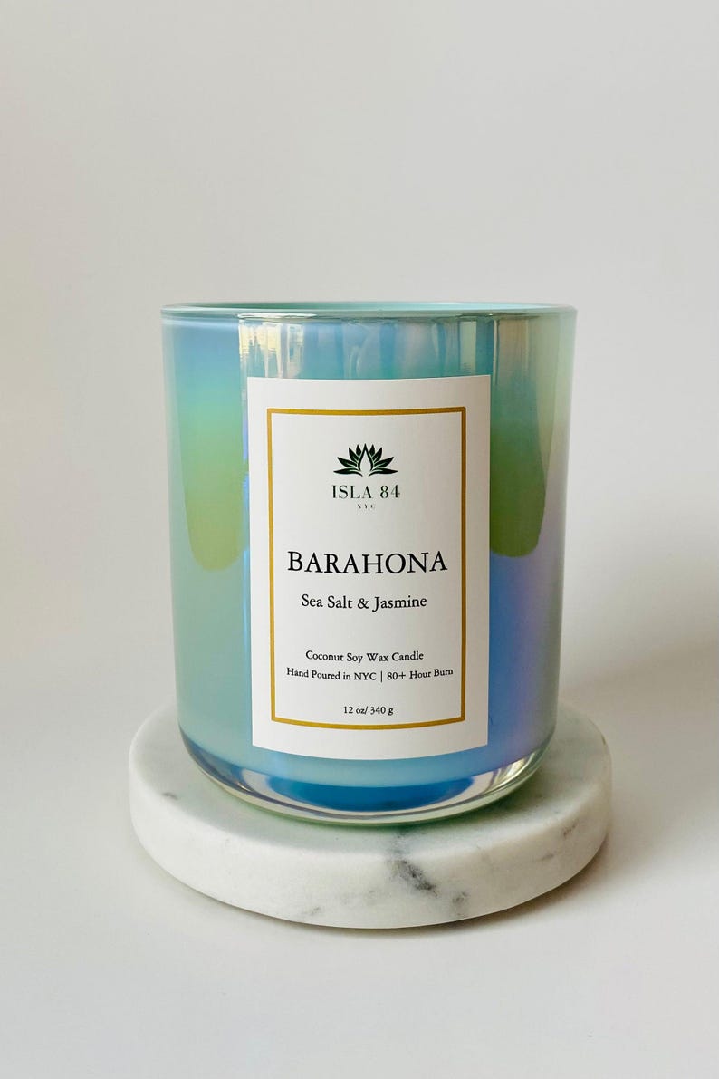 Puede incluir: Un tarro de vela de vidrio iridiscente azul verdoso con una etiqueta blanca que dice "ISLA 84 NYC" y "BARAHONA Sea Salt & Jasmine". La etiqueta tambi&eacute;n incluye el texto "Coconut Soy Wax Candle", "Hand Poured in NYC", "80+ Hour Burn", y "12 oz/340 g".