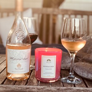 K&ouml;nnte beinhalten: Eine Flasche Whispering Angel Ros&eacute;wein, eine rosa Kerze und zwei Weingl&auml;ser auf einem Holztisch. Die Kerze ist mit "Punta Cana" beschriftet und die Weinflasche hat ein dekoratives Etikett. Die Weingl&auml;ser sind mit Ros&eacute;wein gef&uuml;llt.