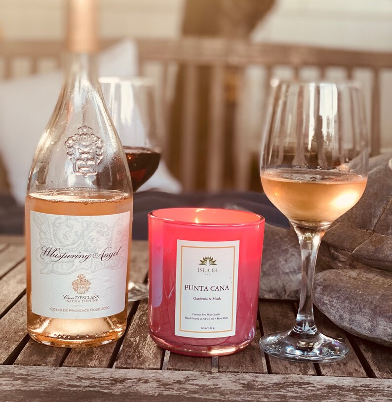 K&ouml;nnte beinhalten: Eine Flasche Whispering Angel Ros&eacute;wein, eine rosa Kerze und zwei Weingl&auml;ser auf einem Holztisch. Die Kerze ist mit "Punta Cana" beschriftet und die Weinflasche hat ein dekoratives Etikett. Die Weingl&auml;ser sind mit Ros&eacute;wein gef&uuml;llt.