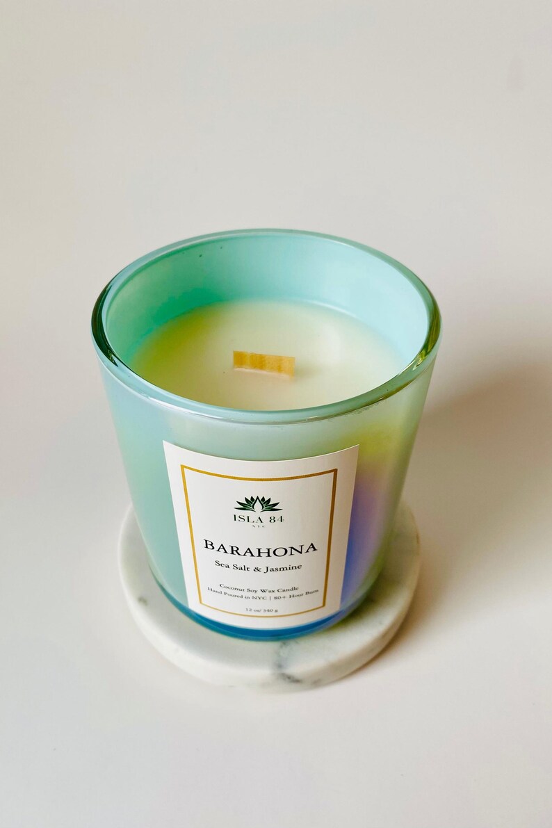 Puede incluir: Un tarro de vela de vidrio azul verdoso con una etiqueta blanca que dice "ISLA 84" y "BARAHONA Sea Salt & Jasmine". La vela est&aacute; hecha de cera de soja y coco y tiene una mecha de madera.